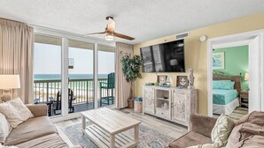 A spacious living room with a large sliding glass door leading to a balcony overlooking the ocean, a ceiling fan, a flat-screen TV, and a cozy seating area with a coffee table. - Pelican Beach 814 — 8th-Floor Oceanfront 2BR Condo in Destin (Destin)