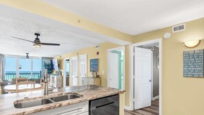 A modern kitchen with a marble countertop, stainless steel sink, and a dishwasher. There is a ceiling fan, a view of the ocean, and a sign listing cottage rules. - Pelican Beach 814 — 8th-Floor Oceanfront 2BR Condo in Destin (Destin)
