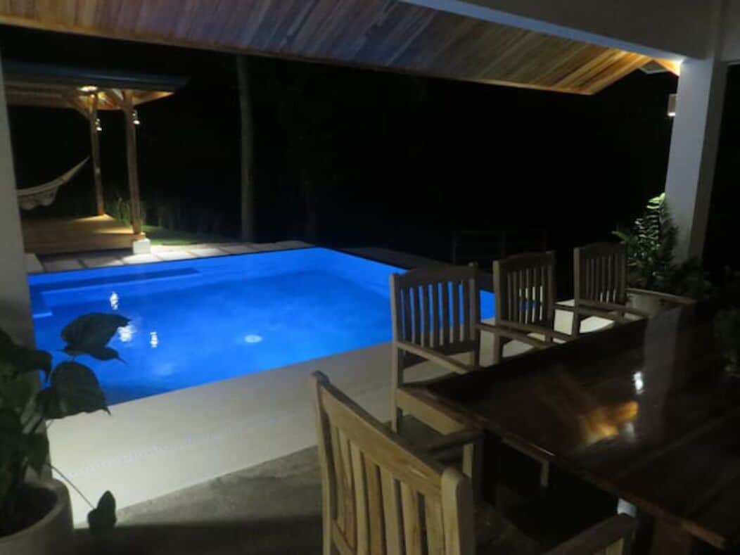 Une piscine dotée d’une terrasse en bois et de chaises, entourée par le ciel sombre de la nuit.