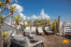 Un salon sur le toit doté de plusieurs espaces de détente, comprenant des fauteuils et des canapés confortables, le tout agrémenté de plantes en pot et de palmiers.