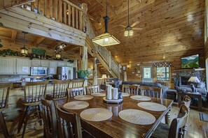 A spacious wooden cabin interior with a dining area, kitchen, living room, and a staircase leading to an upper level.