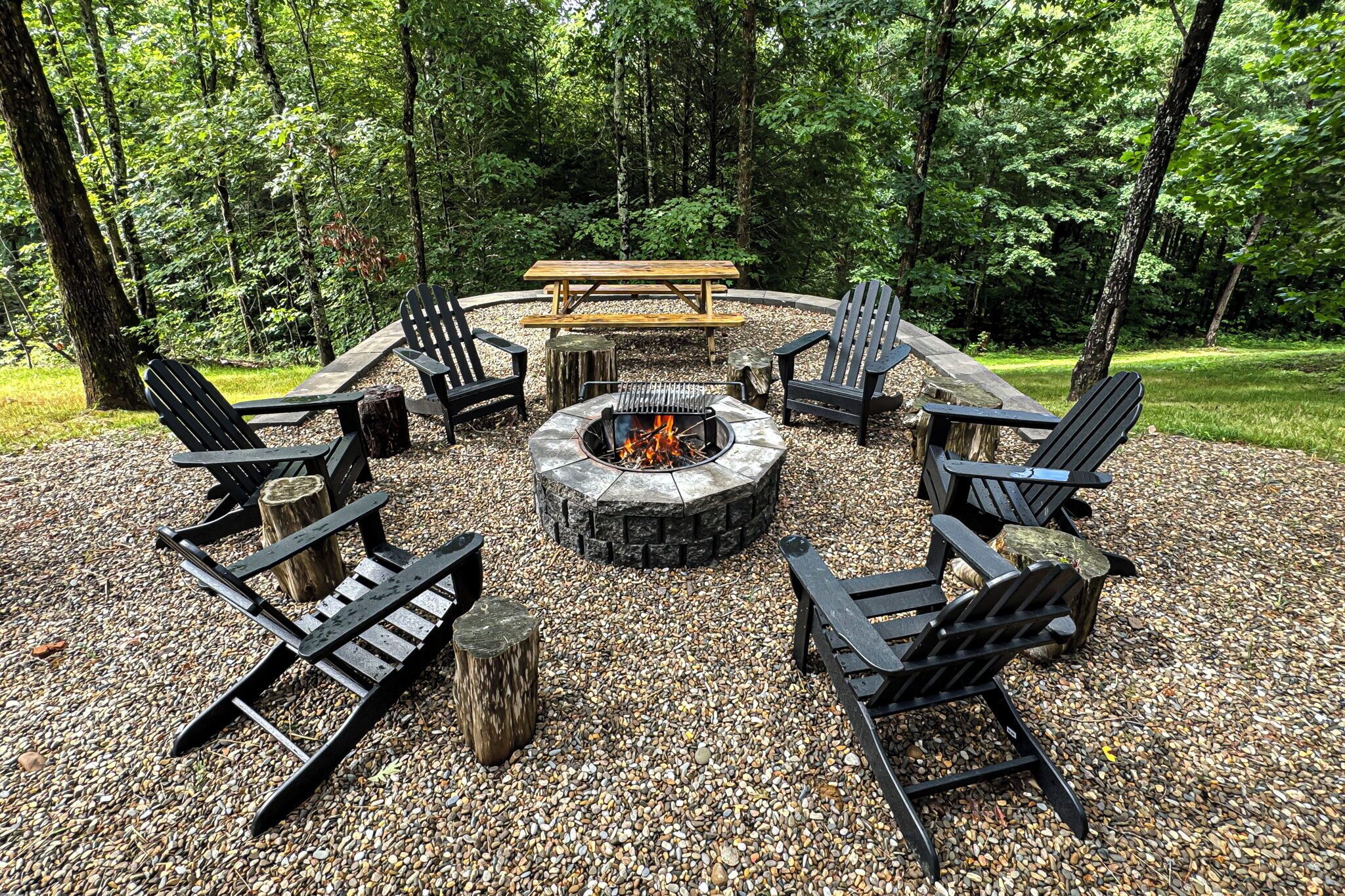 A circular outdoor seating area with a fire pit, surrounded by Adirondack chairs and a picnic table, set on a gravel surface with trees in the background.
