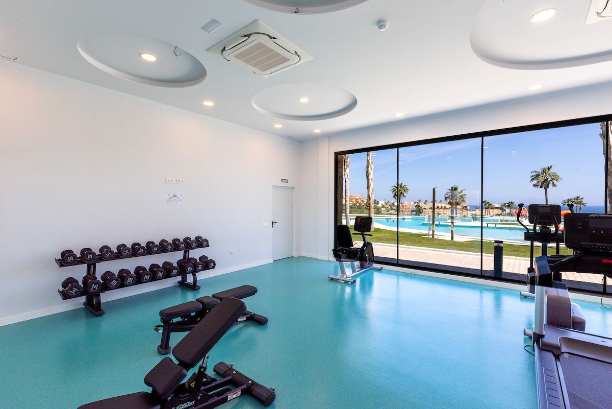 Une salle de sport équipée de matériel d’exercice, d’un banc, d’haltères et offrant une vue sur une piscine et des palmiers.