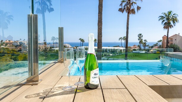 Une table en verre avec une bouteille de vin et des verres, donnant sur une piscine bordée de palmiers et offrant une vue sur l’océan.