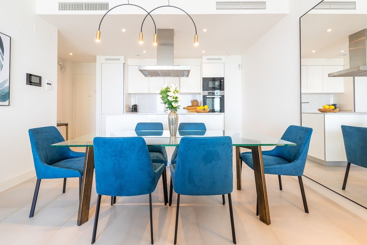A modern dining area with a glass table, blue chairs, and a kitchen in the background.