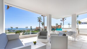 Un espace de détente extérieur moderne, avec des meubles blancs, une balustrade en verre et une vue sur la mer et les palmiers.