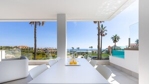 Un espace de restauration extérieur moderne, avec une table et des chaises blanches, offrant une vue sur un paysage côtier agrémenté de palmiers et donnant sur la mer.