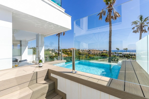 A modern outdoor pool area with glass railings, wooden deck, and a view of the coastline.