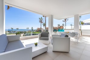 Un área lounge moderna al aire libre con vista al mar y palmeras, con mobiliario blanco y una piscina.
