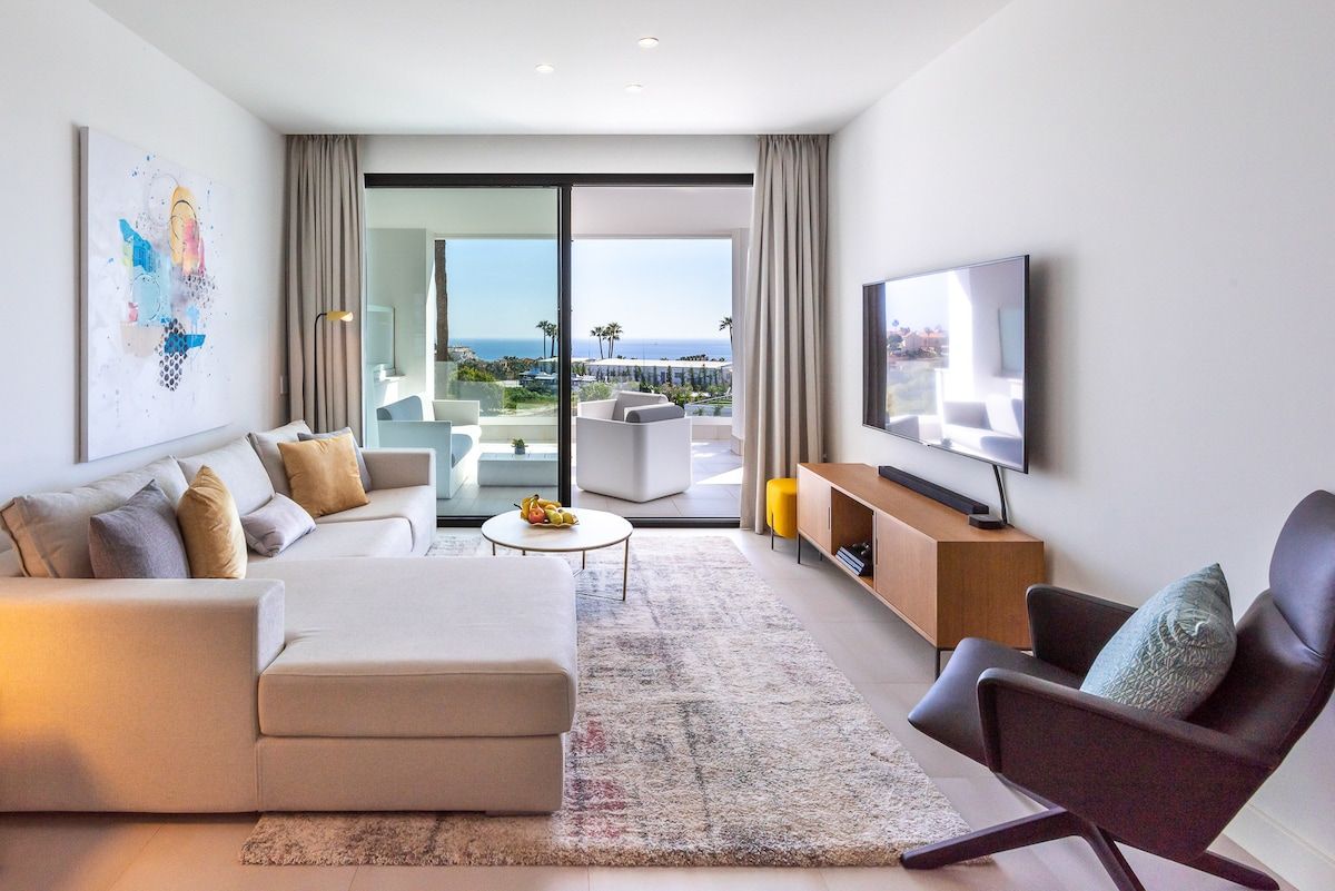Un salon moderne avec un canapé, un fauteuil et un téléviseur à écran plat, une table basse et une vue sur l’extérieur.