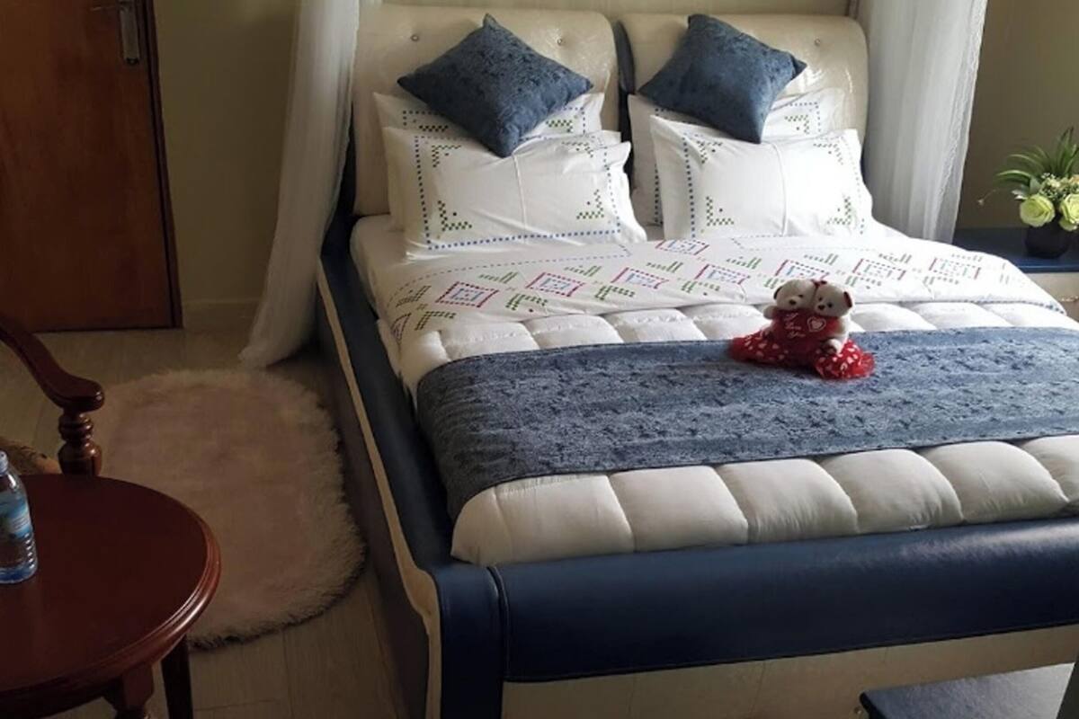 A neatly made bed with a blue and white bedspread, a teddy bear on the bed, a wooden chair, a small round table with a water bottle, and a potted plant on a nightstand.