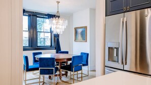 Una cocina moderna con un refrigerador de acero inoxidable, un comedor con mesa de madera y sillas azules, y un ventanal con cortinas azules.