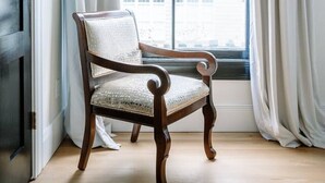 Una silla de madera con asiento y respaldo acolchados, ubicada frente a una ventana con cortinas blancas.