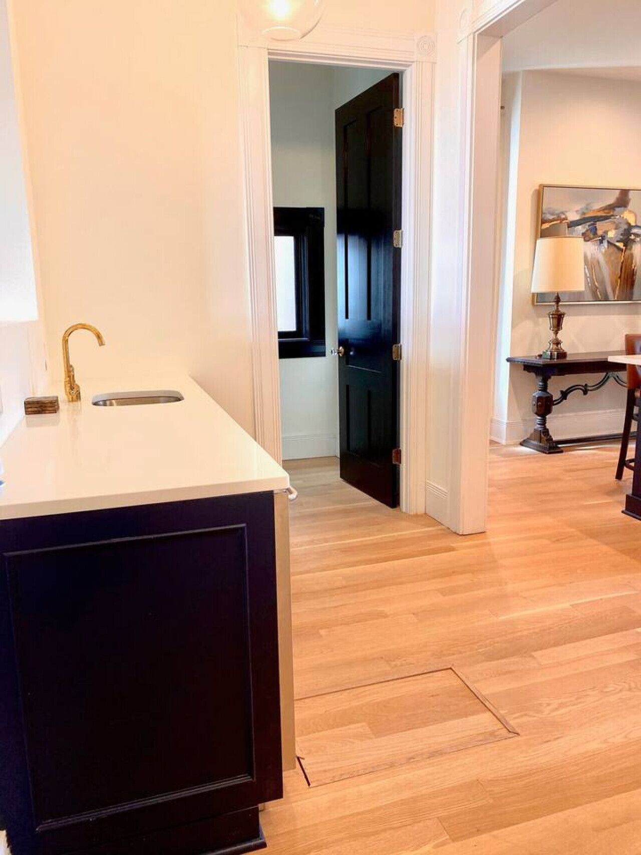 A room with a wooden floor, a dark-colored cabinet, a white countertop with a gold faucet, and a door leading to another room.