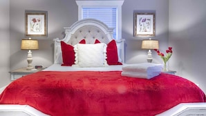 Un dormitorio con una cama grande, ropa de cama roja, un cabecero blanco, dos lámparas de noche y cuadros con motivos florales en las paredes.