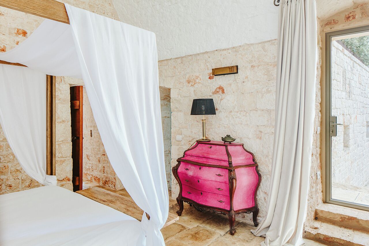 A bedroom with a four-poster bed, a pink dresser, a lamp, and a stone wall.