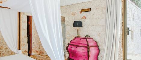 A bedroom with a four-poster bed, a pink dresser, a lamp, and a stone wall.