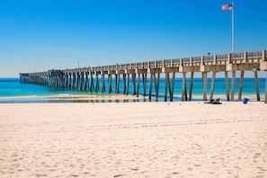 A pier extending into the ocean with a flag at the end, a sandy beach, and a clear sky. - Mariner by iTrip Panama City Beach (Panama City Beach)