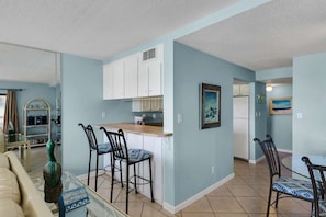 A living room with a beige sofa, a coffee table, and a decorative sculpture. There is a kitchen area with white cabinets, a countertop, and a refrigerator. - Mariner by iTrip Panama City Beach (Panama City Beach)