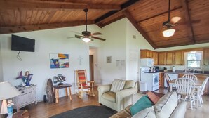 A spacious living area with a sofa, a rocking chair, a dining table with chairs, a kitchen with wooden cabinets, and a ceiling fan.