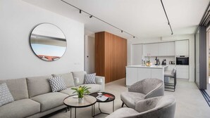 A modern living room with a sofa, armchairs, a coffee table, and a round mirror. A kitchen with white cabinets and a dining area is visible in the background.