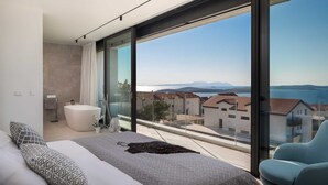 A modern bedroom with a large bed, a bathtub, and a view of a coastal town.