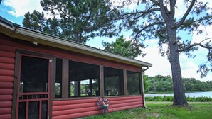 A log cabin with a large windowed porch, a small outdoor seating area, and a view of a lake.