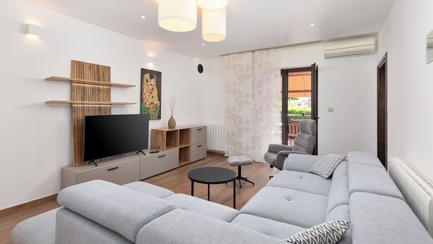 A modern living room with a large flat-screen TV, grey sofas, a small round table, and a wooden shelving unit. - Apartment Mares Family 5+ 100m2 with parking and garden (Porec)