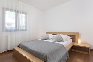 Une chambre à coucher comprenant un lit, des tables de chevet, une commode, un banc, une fenêtre avec des rideaux légers et un radiateur.