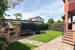Un jardin avec une terrasse en pierre, un barbecue, une table à manger avec des chaises et un panier de basketball.