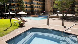 Et rektangulært swimmingpool med flisekant, omgivet af en murstensplads. Loungestole og parasoller ses i baggrunden, og en bygning med flere etager og altaner.