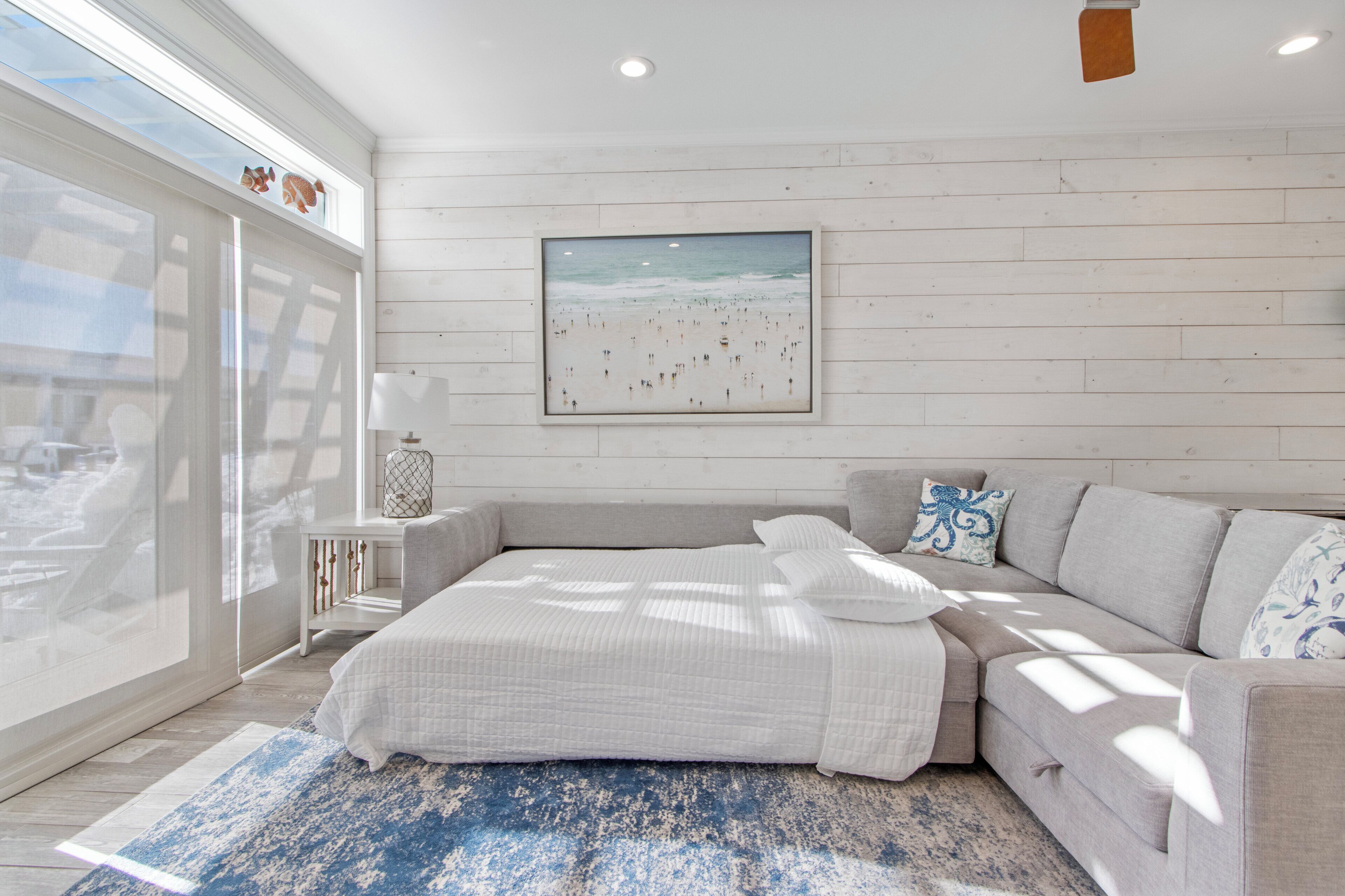 Een slaapkamer met een groot bed, uitzicht op het strand, een lijst met een afbeelding aan de muur, en een klein nachtkastje met een lamp.