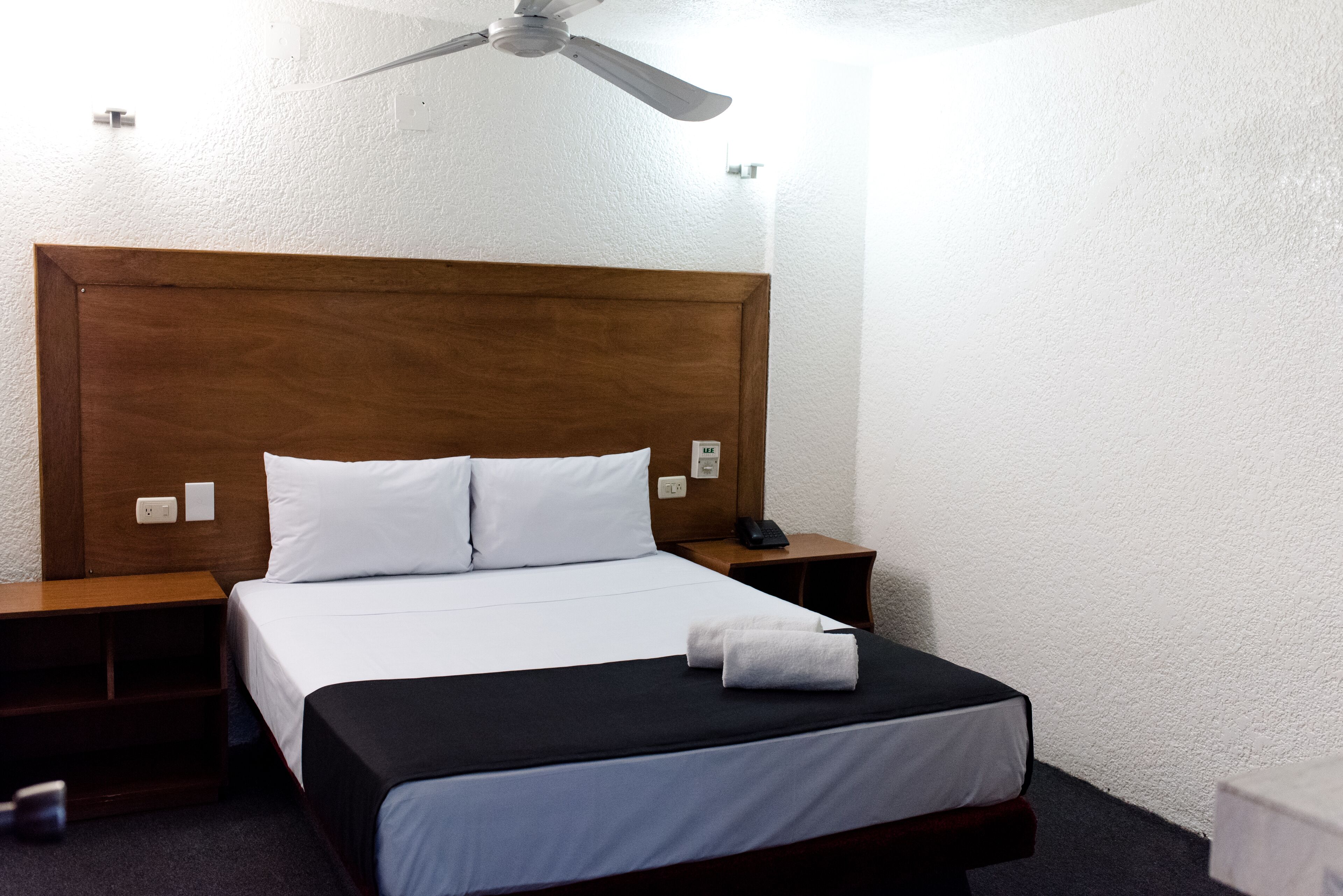 A hotel room with a wooden headboard, a bed with white linens, a black bedspread, and a ceiling fan.
