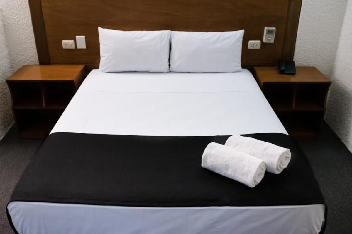 Una habitación de hotel con una cama, dos mesitas de noche, una colcha blanco y negro y dos toallas enrolladas sobre la cama.