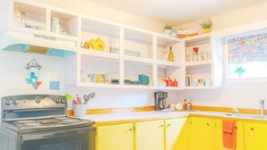 Een keuken met gele kastjes, een fornuis en diverse keukenspullen op planken.