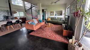 A modern living room with a sofa, coffee table, and a TV. There is a staircase leading to another level, and a dining area with chairs and a table.