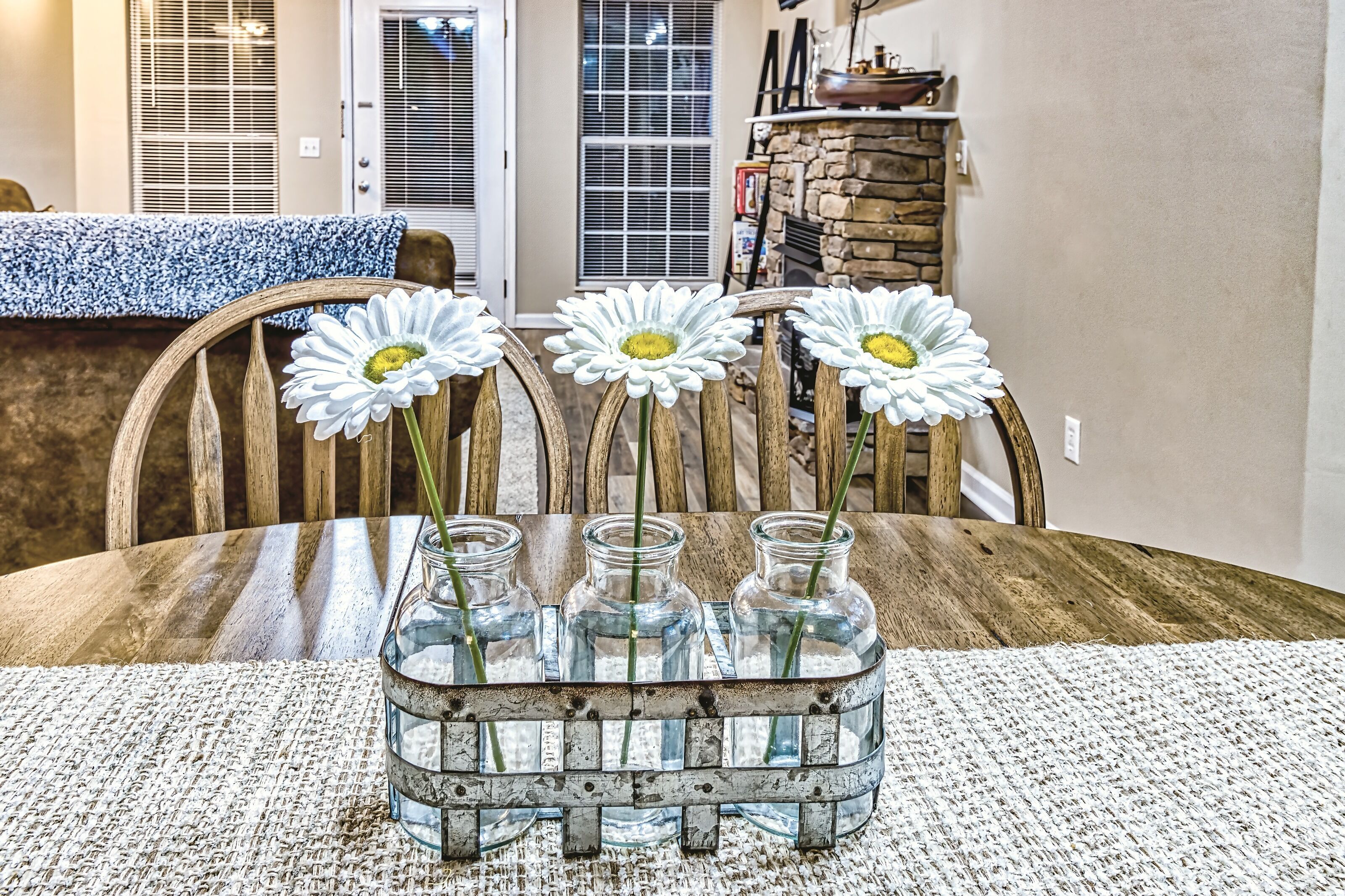 A wooden dining table with a woven placemat, three glass vases with daisy flowers, and a rustic metal tray.