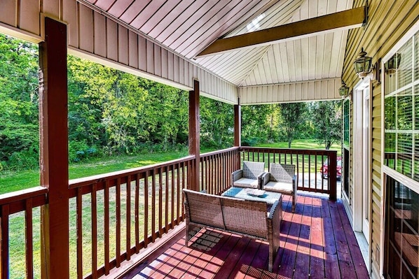 A covered porch with a wooden deck, wicker furniture, and a view of a green landscape.