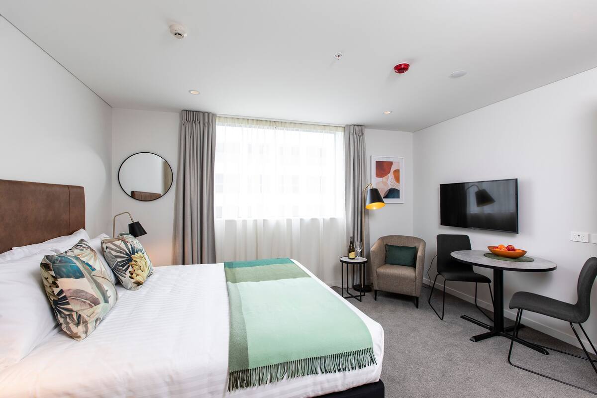 Une chambre d’hôtel avec un lit, un miroir rond, une télévision, une petite table sur laquelle se trouve un saladier rempli de fruits, et une chaise.