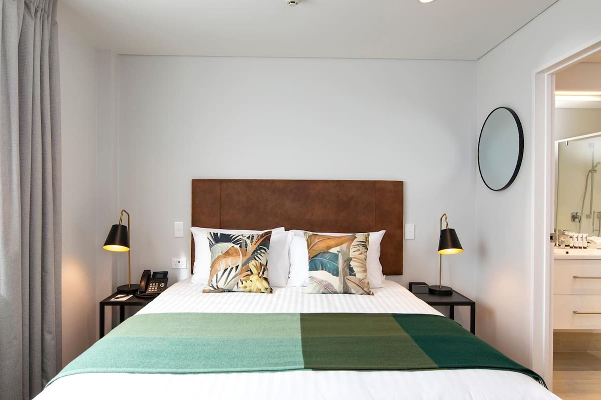 Une chambre d’hôtel avec un lit, deux tables de chevet, un téléphone, un miroir et une salle de bain visible à travers une porte ouverte.