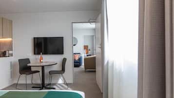 Habitación de hotel con cama, televisor, mesa con dos sillas, cafetera y vista al pasillo.