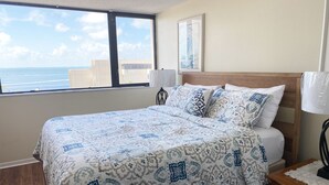 A bedroom with a large bed, a nightstand, a lamp, and a view of the ocean through a window. - Top floor condo in Key Largo with spectacular Sunset views (Key Largo)