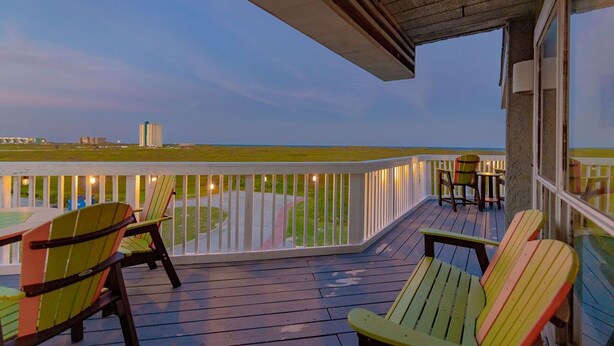 A wooden deck with Adirondack chairs and a table, overlooking a grassy area and the ocean. - ⭐⭐⭐⭐⭐ BEAUTIFUL OCEAN VIEWS + Huge Deck + Heated Pool + Hot tub + Boardwalk  (Port Aransas)