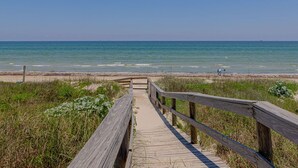 A wooden boardwalk leading to a beach with clear blue water and a distant view of the ocean. - ⭐⭐⭐⭐⭐ BEAUTIFUL OCEAN VIEWS + Huge Deck + Heated Pool + Hot tub + Boardwalk  (Port Aransas)