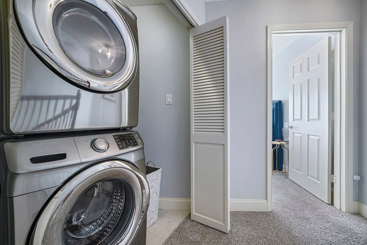 A laundry room with stacked washers and dryers, a white door, and a hallway leading to another room.