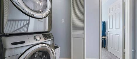 A laundry room with stacked washers and dryers, a white door, and a hallway leading to another room.