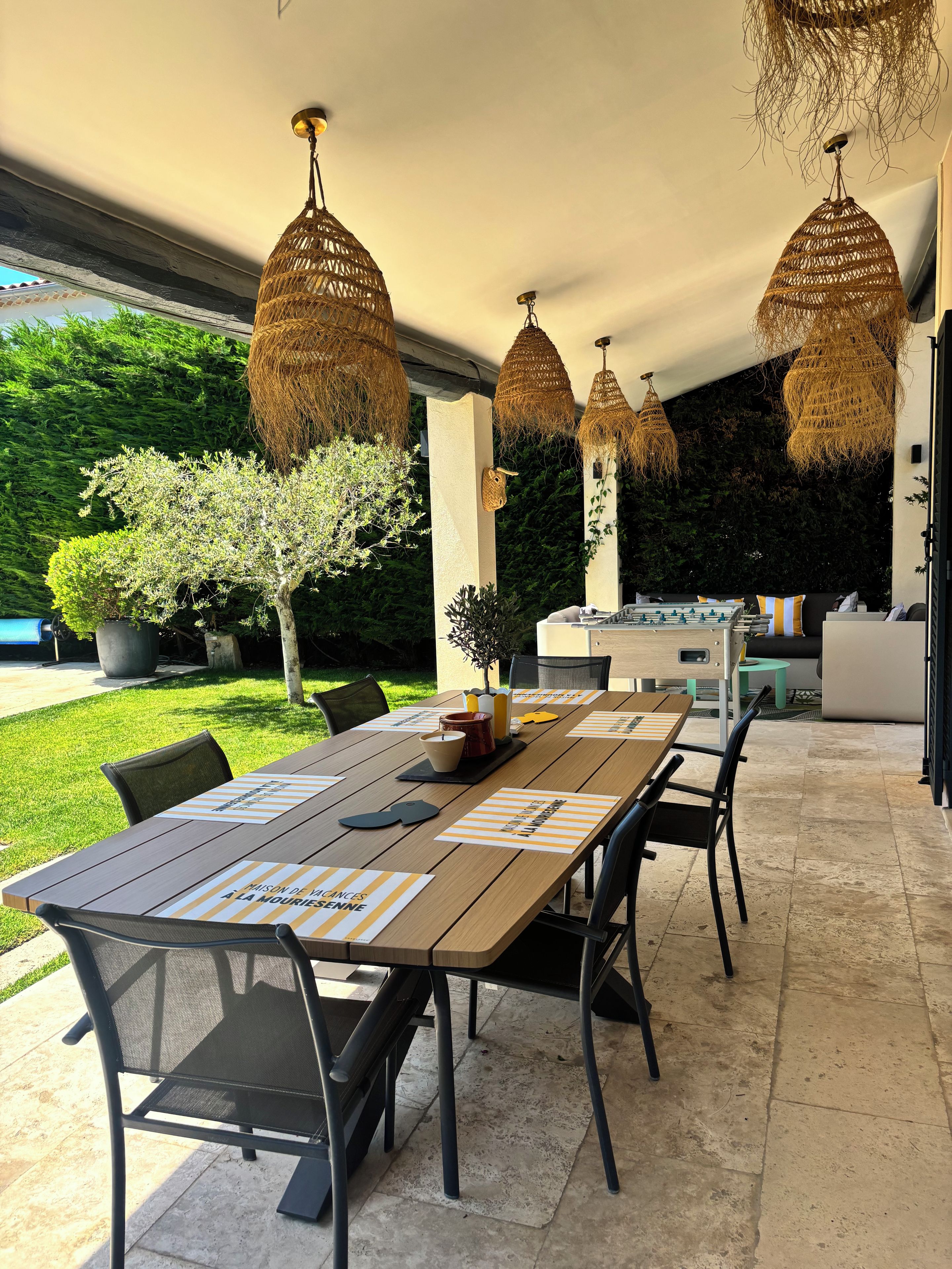 Un espace de restauration en plein air avec une longue table, des chaises et des luminaires suspendus en osier.