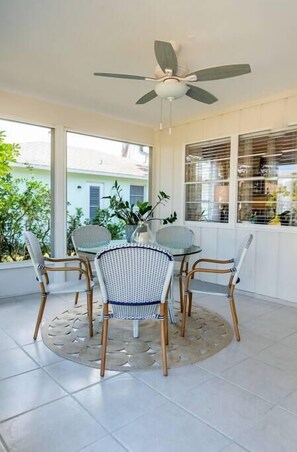 A covered patio with a table and chairs, a ceiling fan, and large windows overlooking a garden.