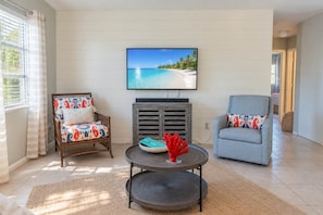 A living room with a television mounted on the wall, two chairs with patterned cushions, a round coffee table with a decorative item, and a view of a beach through the window.