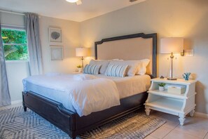 A bedroom with a dark wooden bed, white bedding, two bedside lamps, a white nightstand, and a view of greenery outside.